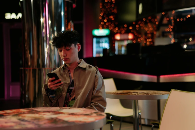 a man sitting at a table looking at his cell phone, by Nick Fudge, pexels, realism, finn wolfhard, multicoloured, standing in a restaurant, evening time