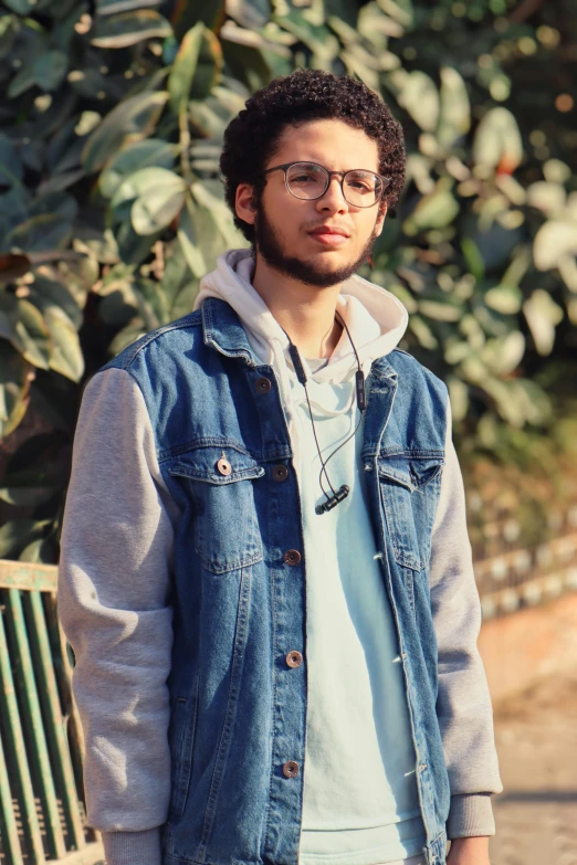 a man standing next to a bench in a park, an album cover, by Ahmed Yacoubi, unsplash, wearing a jeans jackets, jewish young man with glasses, ((portrait)), hooded