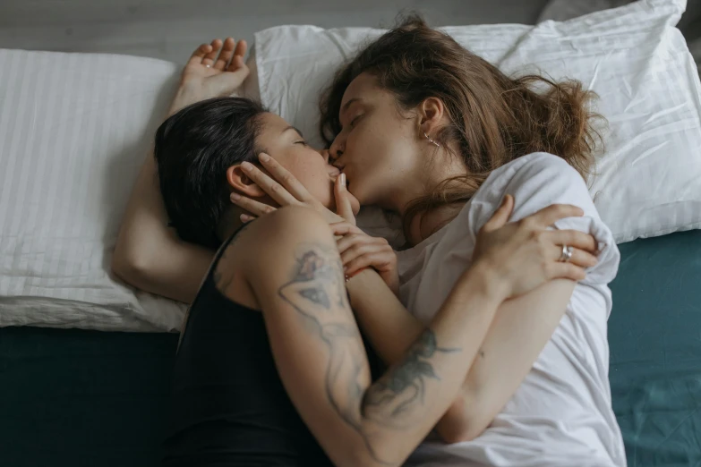 a couple of women laying on top of a bed, trending on pexels, romanticism, lesbian kiss, tattoos and piercings, medium shot of two characters, semi - realistic