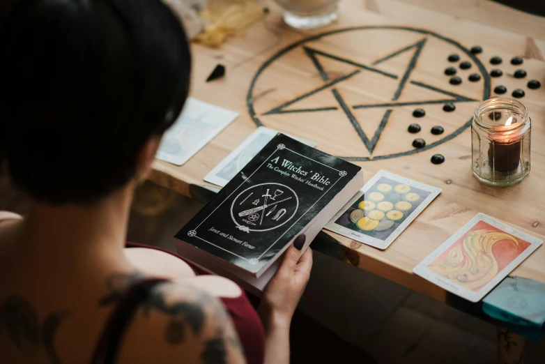 a woman sitting at a table holding a book, pexels contest winner, process art, pentacle, character sheets on table, summoning circle, instagram story