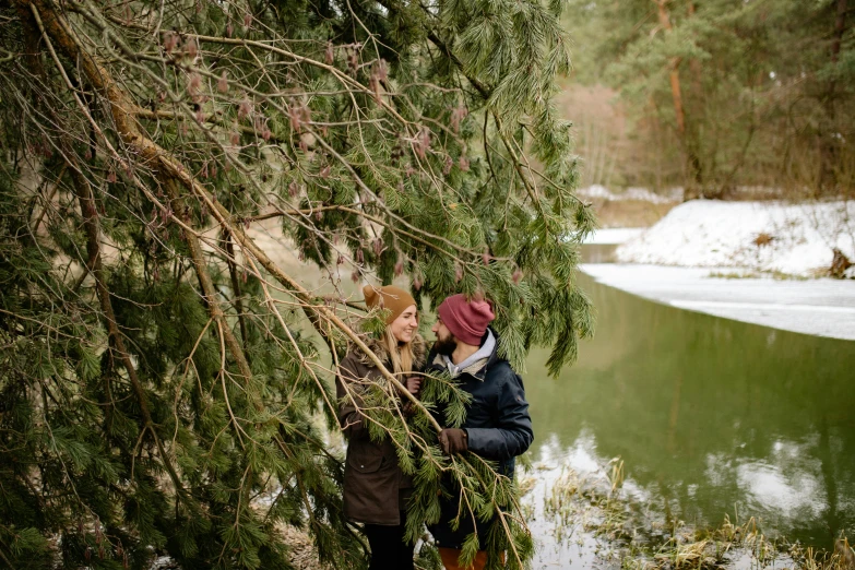 a man and a woman standing next to a body of water, by Julia Pishtar, pexels contest winner, evergreen branches, outside winter landscape, digging, overhanging branches