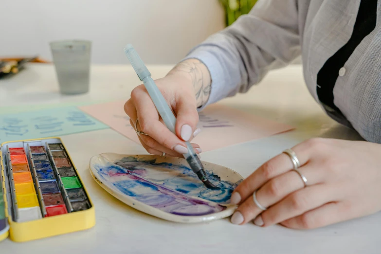 a woman sitting at a table painting with watercolors, trending on pexels, painting on a badge, clay art, animation still, holding pencil