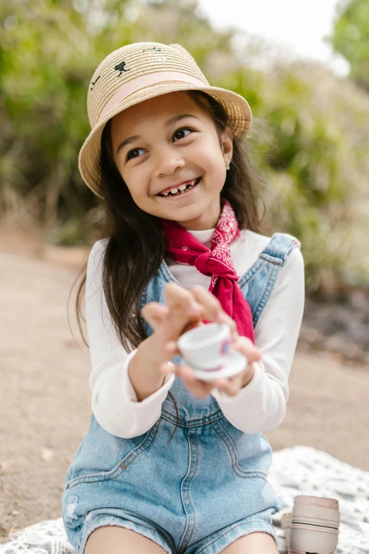 a little girl sitting on top of a blanket, holding a boba milky oolong tea, wearing a travel hat, mini model, detailed product image