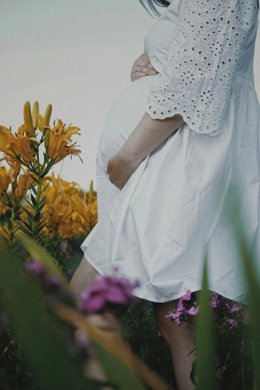 a woman in a white dress standing in a field of flowers, pexels contest winner, symbolism, pregnant belly, next to a plant, close-up on legs, ad image