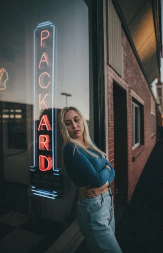 a woman standing in front of a building with a neon sign, by Drew Tucker, pexels contest winner, blonde girl, backroom background, haggard, slightly smirking