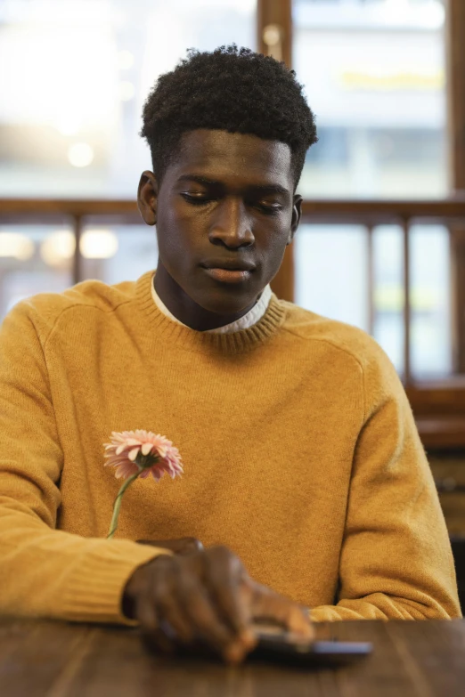 a man sitting at a table with a flower in his hand, by James Morris, trending on unsplash, brown sweater, black teenage boy, ochre, model pose