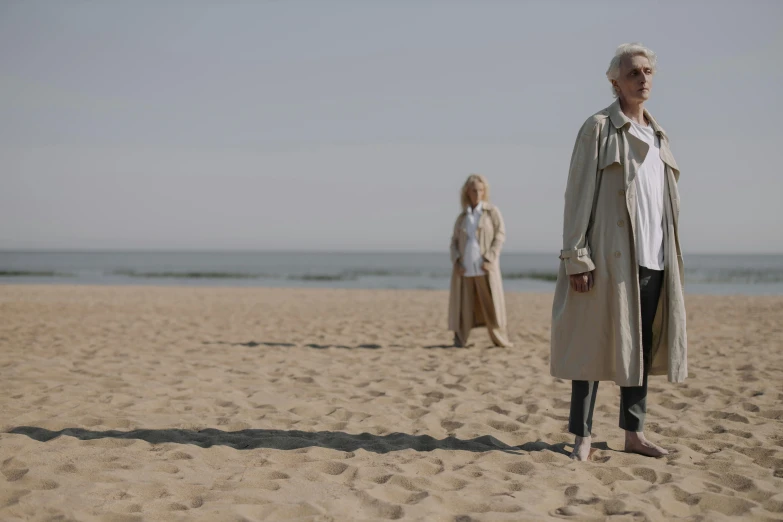 a couple of people standing on top of a sandy beach, unsplash, hyperrealism, woman is in a trenchcoat, white haired lady, ( ( theatrical ) ), midsommar