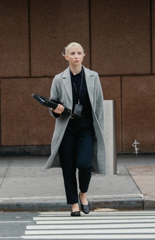 a woman walking across a street next to a crosswalk, pexels, saoirse ronan, grey pants and black dress shoes, carrying a tray, wearing black overcoat