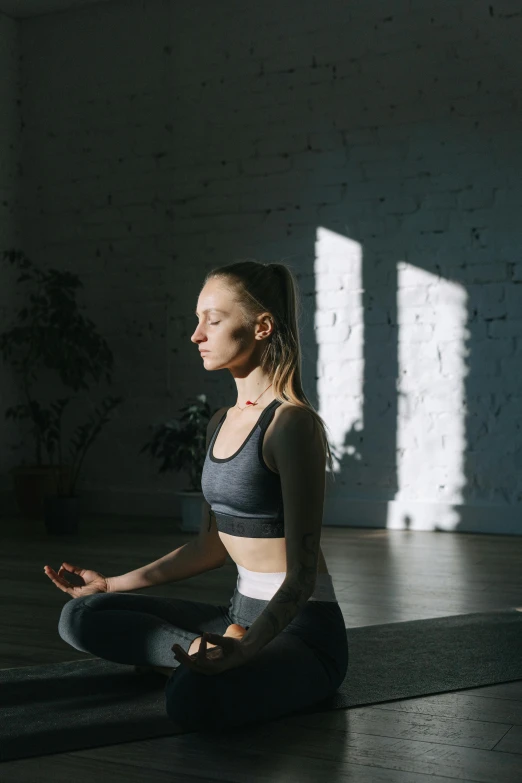 a woman sitting on a yoga mat in a dark room, light and space, slightly sunny, wearing a track suit, meditating, unsplash transparent