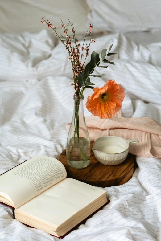 an open book sitting on top of a bed next to a vase of flowers, wearing white pajamas, laying in bed