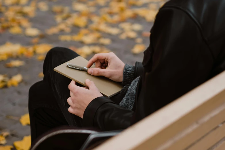 a person sitting on a bench writing on a book, trending on pexels, autumnal, holding a clipboard, a man wearing a black jacket, thin soft hand holding cigarette