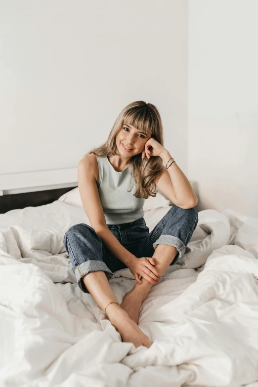 a woman sitting on top of a bed covered in white sheets, trending on pexels, happening, wearing jeans, center parted bangs, blonde swedish woman, avatar image
