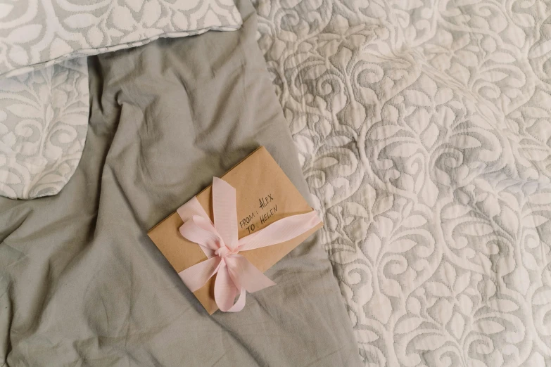 a box sitting on top of a bed next to a pillow, by Alice Mason, pexels contest winner, ribbon, greeting card, grey, pink