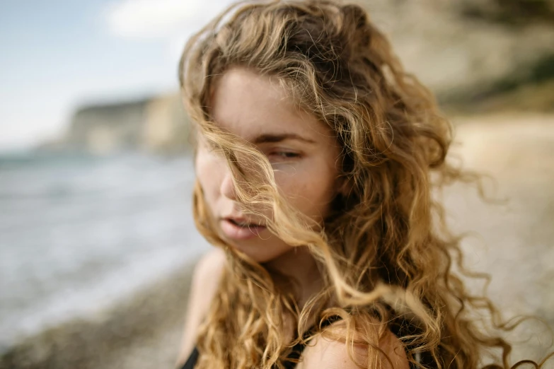 a woman standing on top of a beach next to the ocean, trending on pexels, renaissance, light brown messy hair, exhausted face close up, portrait of teenage aphrodite, hair blowing