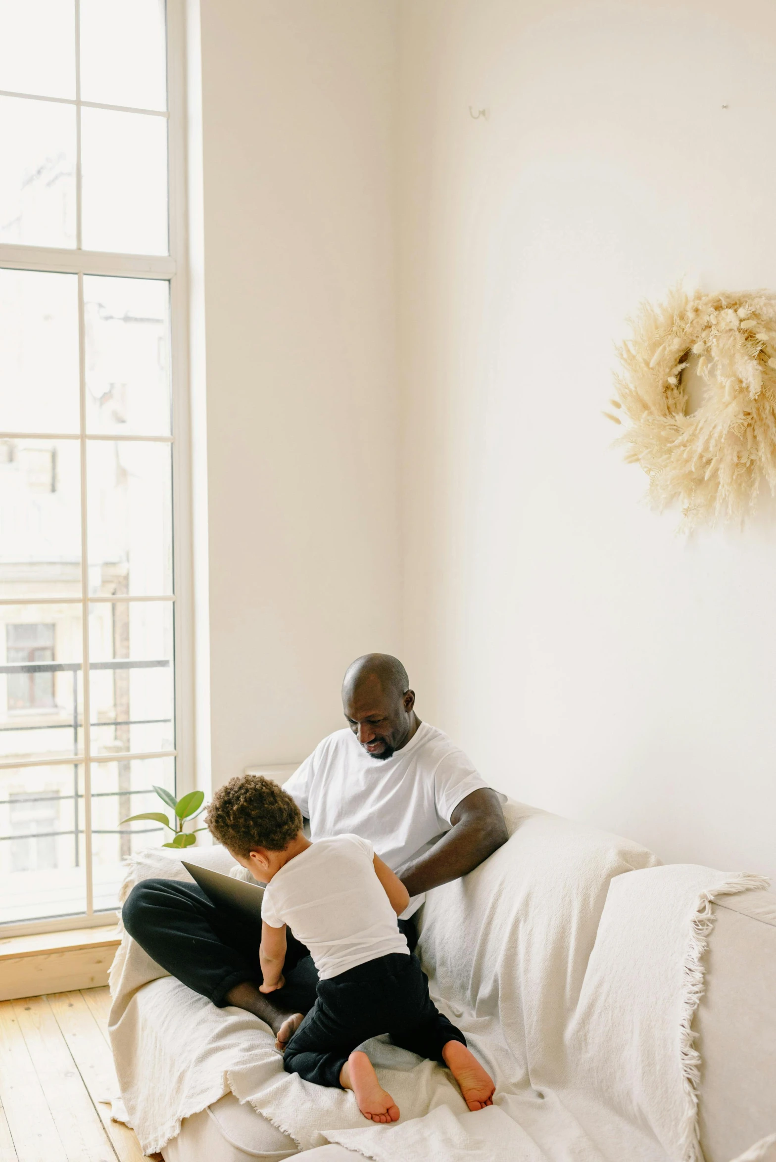 a man sitting on top of a white couch next to a little boy, pexels contest winner, visual art, black man, light beige pillows, comforting, next to window