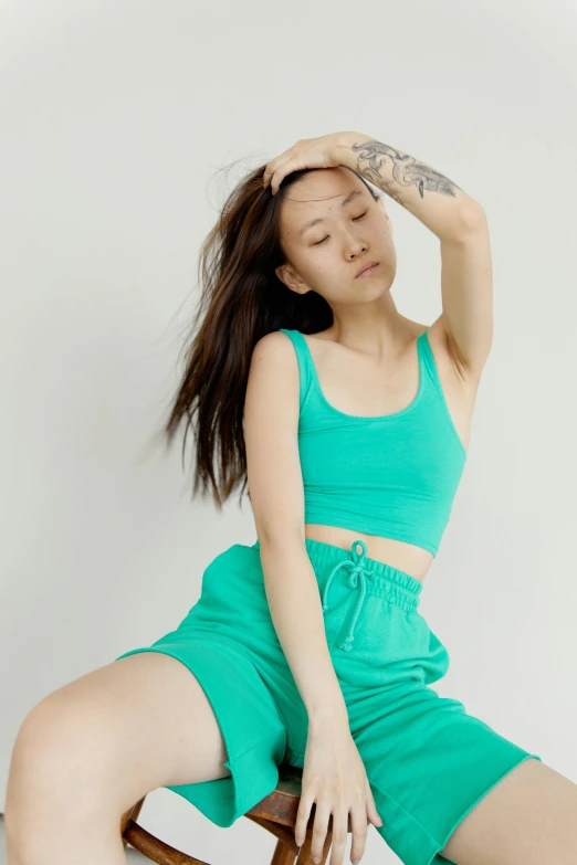 a woman sitting on top of a wooden chair, inspired by helen huang, trending on pexels, wearing green clothing, croptop and shorts, wearing a track suit, cyan