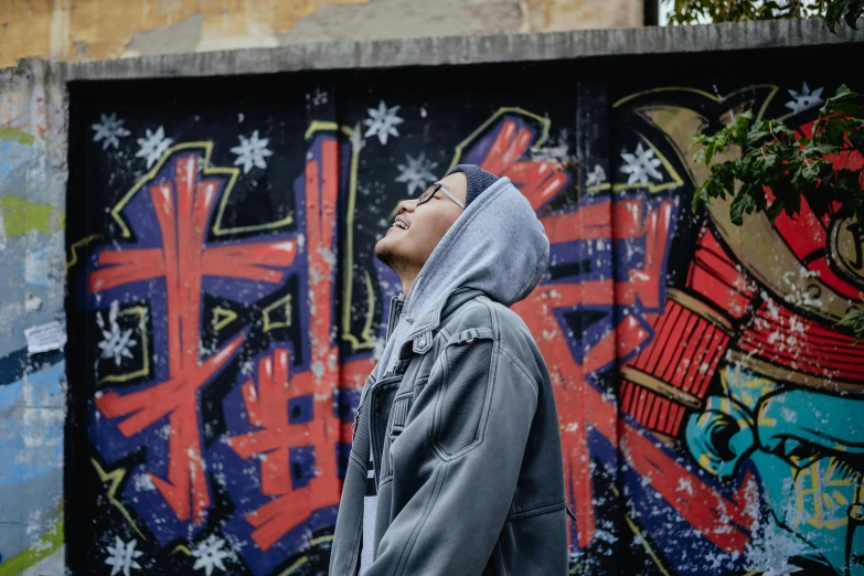 a man standing in front of a graffiti covered wall, inspired by Gang Hui-an, trending on unsplash, graffiti, grey hoodie, looking to the sky, ashteroth, looking happy