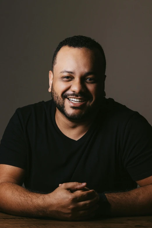 a man sitting at a table smiling at the camera, an album cover, inspired by Samuel Silva, headshot profile picture, dark backdrop, giorgio a. tsoukalos, arab ameera al-taweel