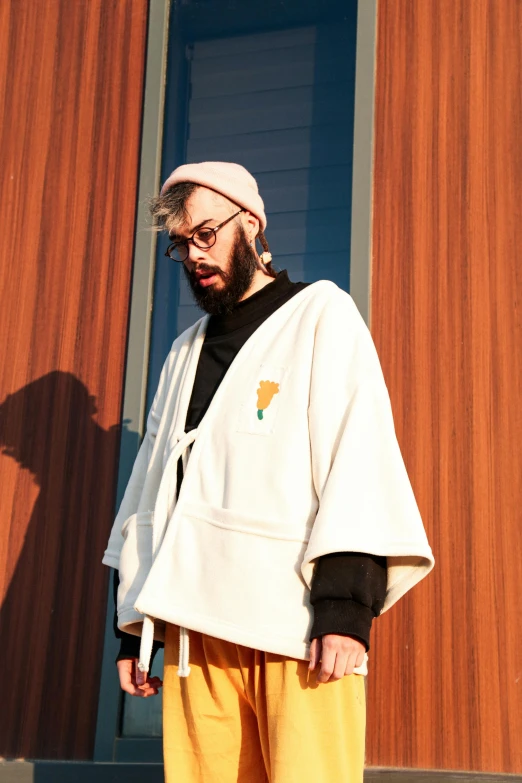 a man standing in front of a building, an album cover, by Bascove, unsplash, neo-dada, wearing white robe, non-binary, beard, cardigan