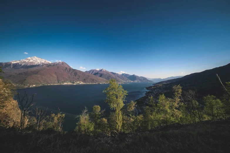 a large body of water surrounded by trees, by Daniel Lieske, pexels contest winner, the alps are in the background, boka, blue sky, 4 k cinematic panoramic view