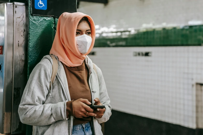 a woman wearing a face mask looking at her phone, trending on pexels, graffiti, muslim, subway station, background image, square