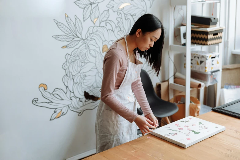 a woman cutting a cake on top of a wooden table, a silk screen, inspired by Qian Xuan, pexels contest winner, wall painting, dressed in a flower dress, drawn on white parchment paper, in a studio