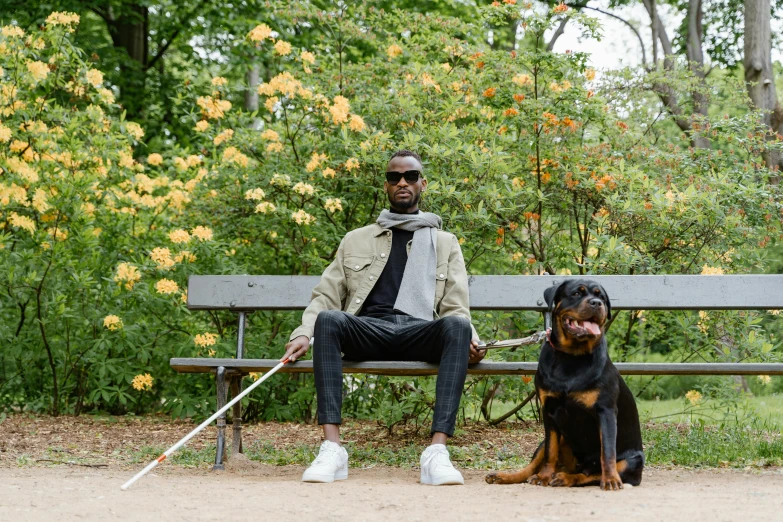 a man sitting on a bench next to a dog, by Daniel Gelon, two crutches near bench, dark-skinned, off - white collection, with a park in the background