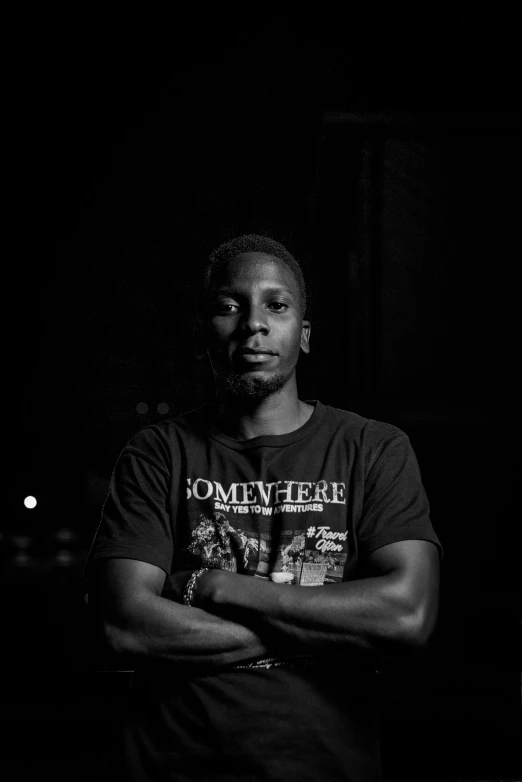 a man standing in the dark with his arms crossed, a black and white photo, by Stokely Webster, he is wearing a black t-shirt, black teenage boy, technical, album