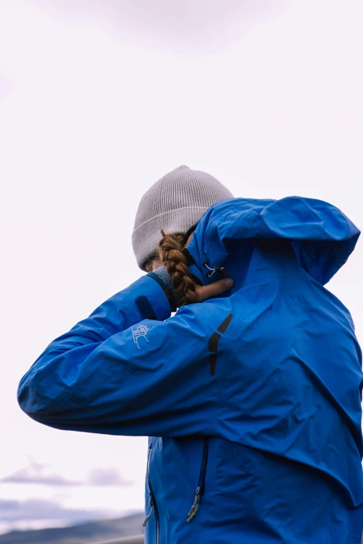 a person in a blue jacket talking on a cell phone, with a covered face, wearing adventuring gear, head bowed slightly, it's raining