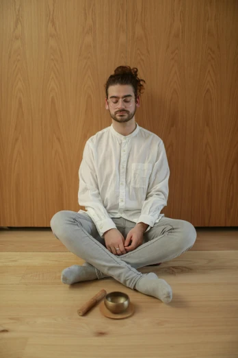 a man sitting on the floor in front of a wooden wall, by Nina Hamnett, unsplash, renaissance, wearing white pajamas, meditating pose, plain background, androgynous person