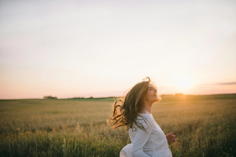 a woman standing in a field at sunset, pexels contest winner, happening, smiling playfully, wind in hair, girl running, facing sideways