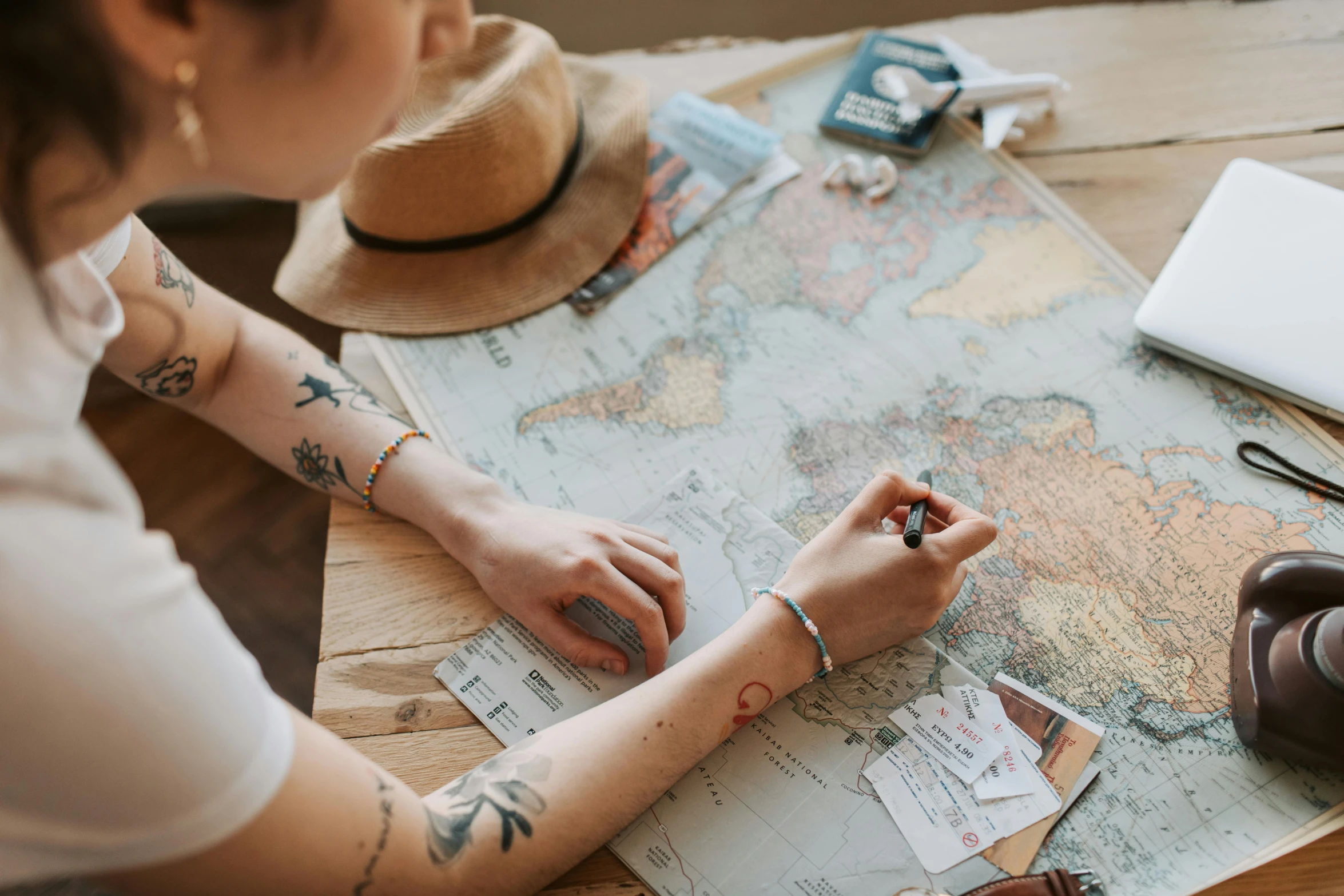 a woman sitting at a table looking at a map, by Emma Andijewska, trending on pexels, with tattoos, crafts and souvenirs, wearing a travel hat, thumbnail