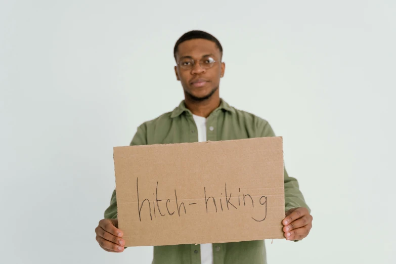 a man holding a sign that says hitch - hiking, trending on pexels, ( ( dark skin ) ), brown, thick lining, in front of white back drop