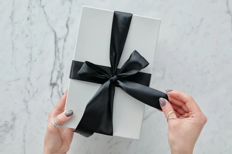 a woman holding a white box with a black bow, inspired by Eden Box, private press, extremely polished, close up shot from the side, holding gift, grey