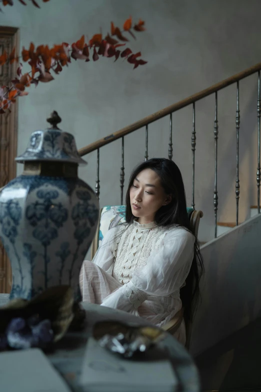 a woman sitting in a chair next to a vase, inspired by Zhou Shuxi, pexels contest winner, thoughtful pose, late autumn, sitting on a grand staircase, medium format. soft light