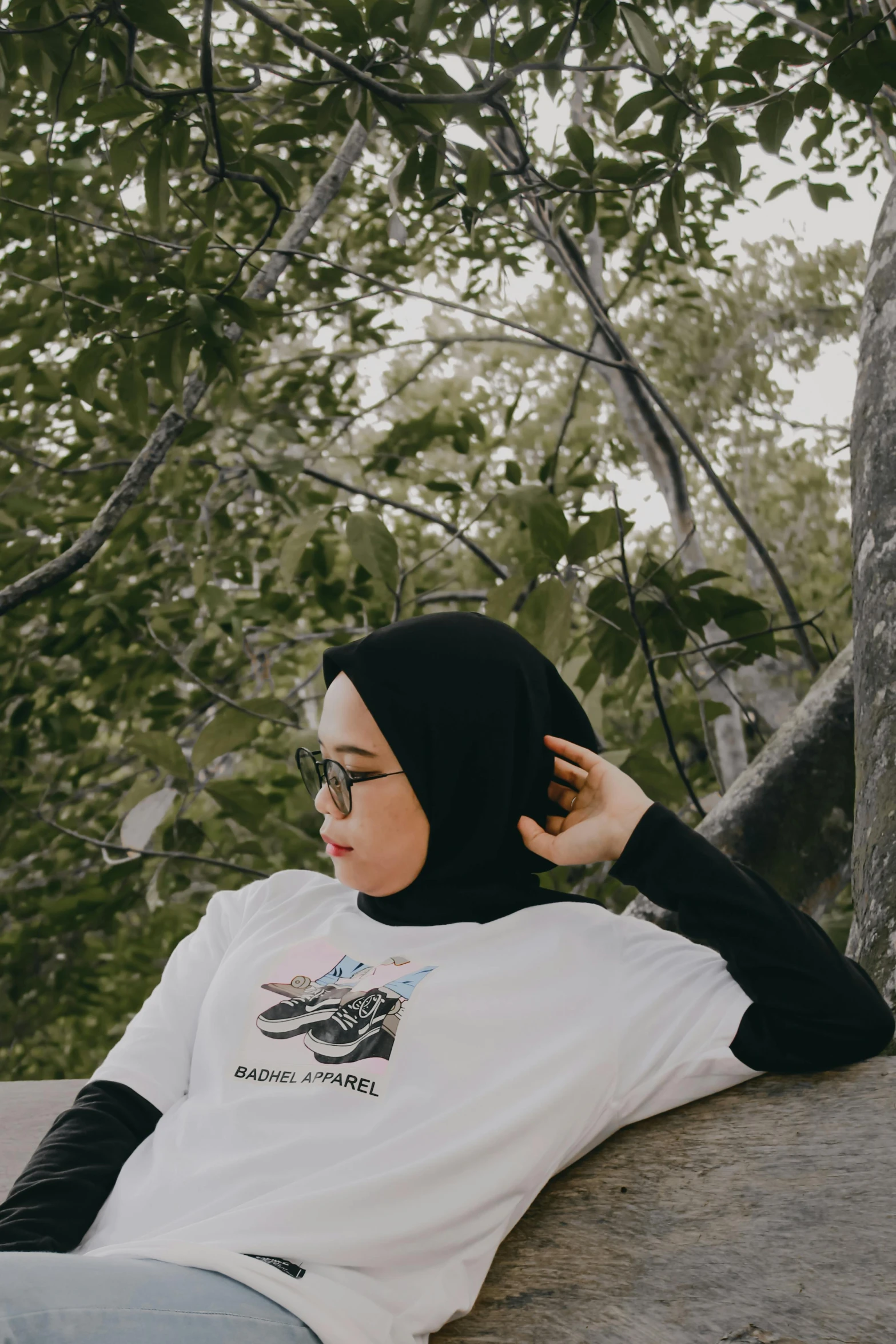 a woman sitting on top of a rock next to a tree, inspired by Naza, realism, plain white tshirt, hijab, wearing black frame glasses, trending on r/streetwear