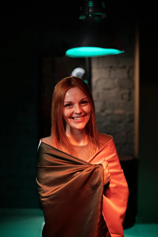 a woman wrapped in a blanket in a dark room, smiling at camera, copper and emerald, in a nightclub, nina tryggvadottir