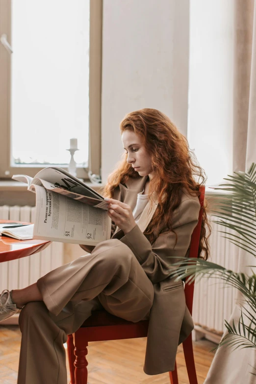 a woman sitting in a chair reading a newspaper, trending on pexels, flowing ginger hair, tan suit, androgynous person, scandinavian