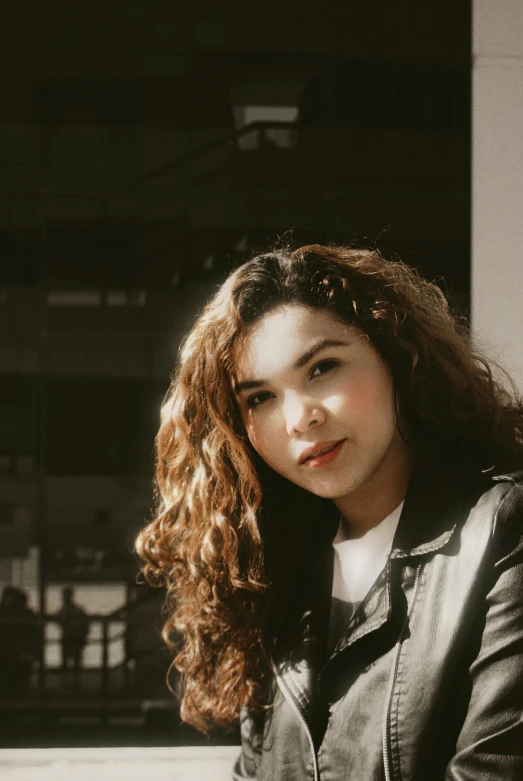 a woman in a leather jacket sitting on a bench, inspired by Elsa Bleda, pexels contest winner, photorealism, her face framed with curls, with backdrop of natural light, ameera al-taweel, grainy low quality photograph