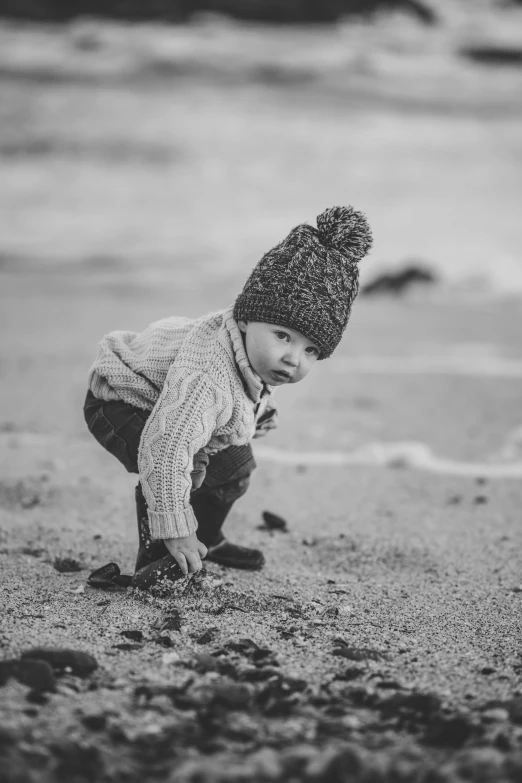 a little boy playing in the sand at the beach, a black and white photo, inspired by Myles Birket Foster, beanie, dressed in a worn, pebbles, looking serious