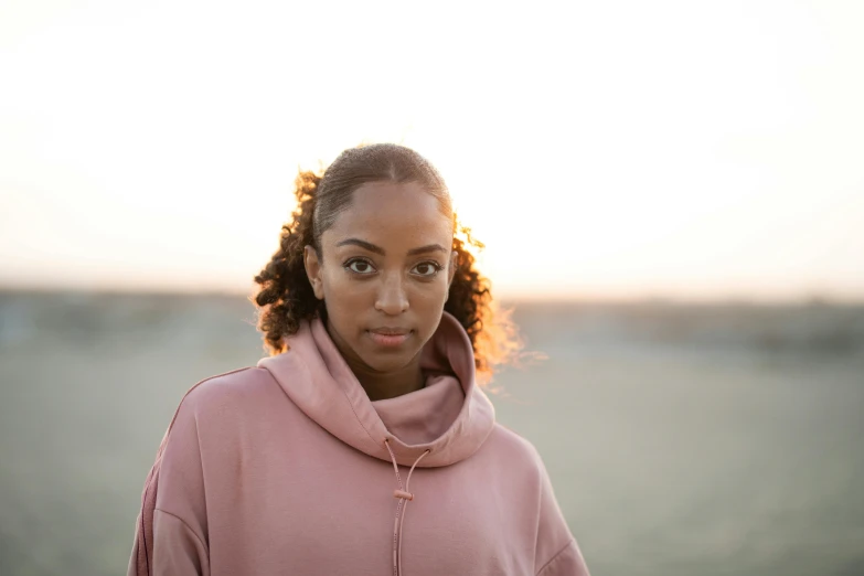 a woman standing on top of a sandy beach, a portrait, trending on pexels, wearing a pastel pink hoodie, ashteroth, focused on her neck, evening light