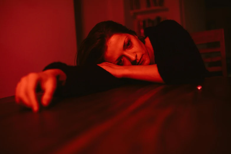 a woman sitting at a table in front of a red light, inspired by Nan Goldin, pexels, portrait of a woman sleeping, sad vampire, on a table, laying on her back