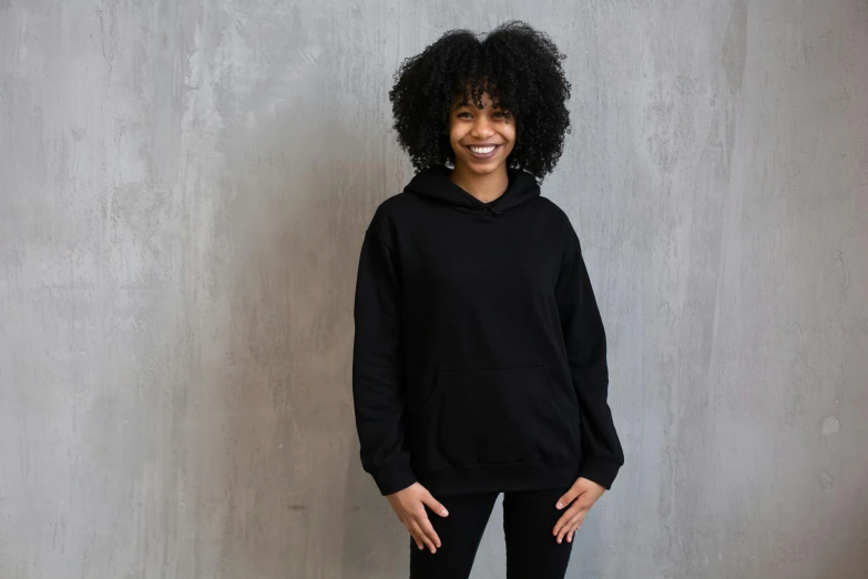 a woman standing in front of a concrete wall, inspired by Mary Black, pexels contest winner, black hoodie, dark short curly hair smiling, main colour - black, thumbnail