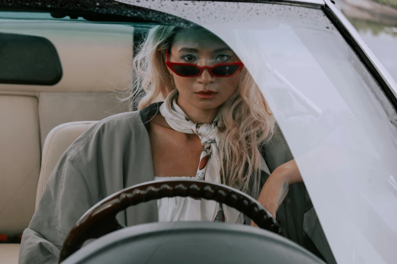 a woman sitting in the driver's seat of a car, inspired by Elsa Bleda, trending on pexels, retrofuturism, wearing torn white cape, wearing versace sunglasses, sydney sweeney, wearing a trenchcoat