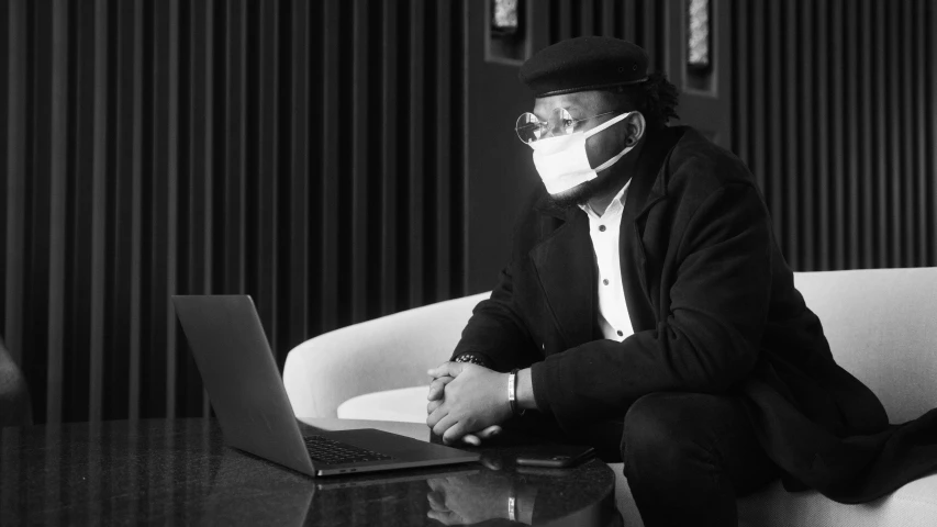 a man sitting on a couch with a laptop, a black and white photo, by Maurycy Gottlieb, pexels, afrofuturism, surgical mask covering mouth, a suited man in a hat, hotei is on the table, worksafe. instagram photo