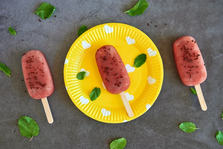 three pops sitting on top of a yellow plate, mint leaves, pepperoni, on grey background, covered with pink marzipan