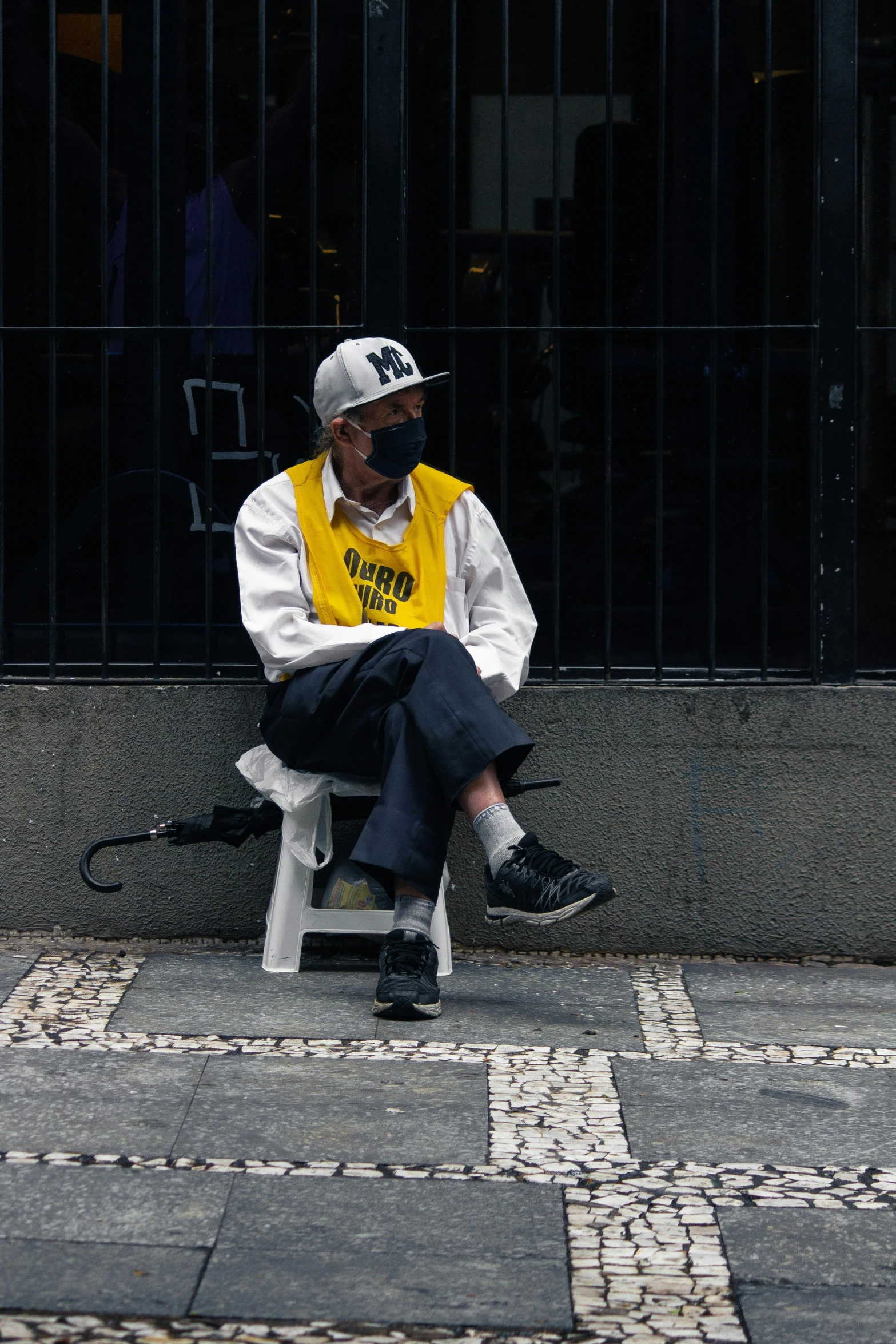 a man sitting on a chair in front of a building, by david rubín, pexels contest winner, street art, gang saints wear yellow bandanas, wearing a vest, white man with black fabric mask, wearing a backwards baseball cap