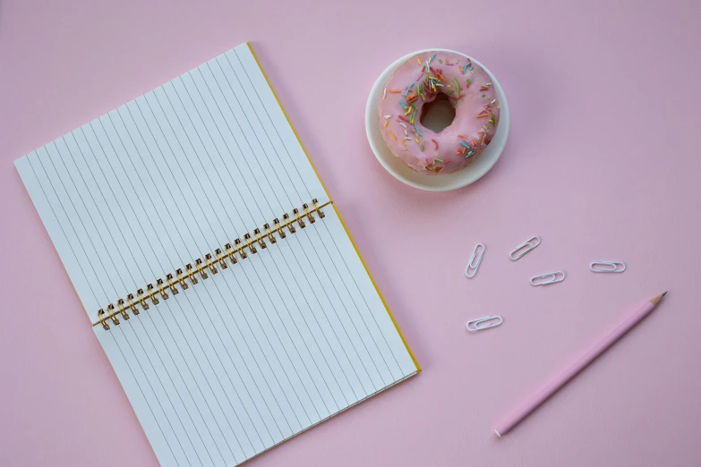 a donut sitting on top of a notepad next to a cup of coffee, trending on pexels, minimalism, pink frosted donut, pencil, background image, unedited