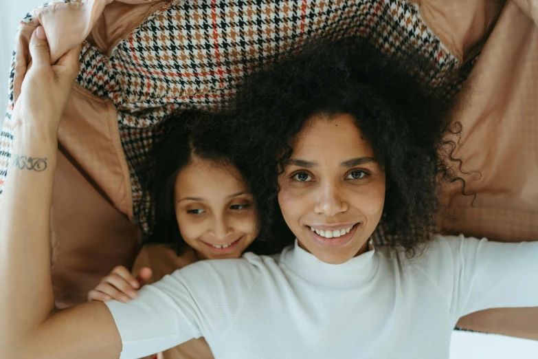 a woman laying on top of a bed next to a little girl, pexels contest winner, she has olive brown skin, smiling down from above, avatar image, curled up under the covers