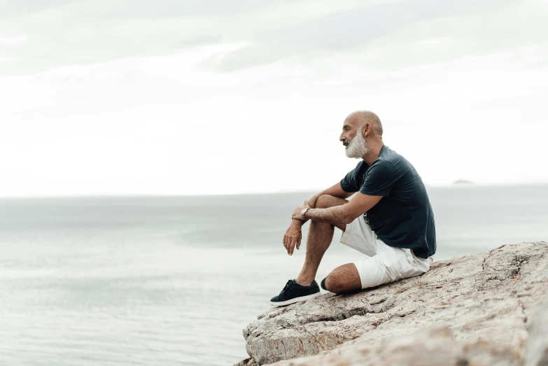 a man sitting on top of a rock next to the ocean, bald head and white beard, wearing shorts and t shirt, profile image, photoshoot for skincare brand